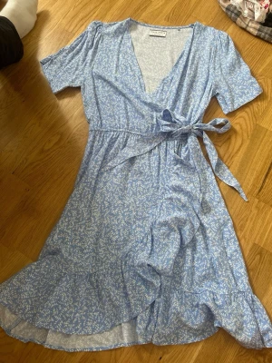 Blå omlottklänning med blommönster - Söt blå kortklänning med vitt blommönster och omlottmodell. Klänningen har v-ringning, volangkant nertill, knytband i midjan och korta ärmar. Perfekt för varma dagar och ger en fräsch sommarkänsla.