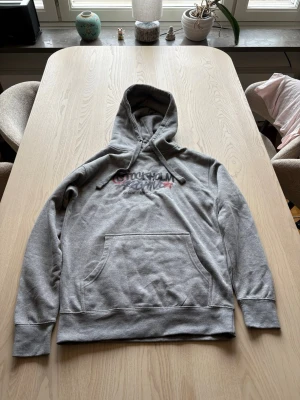 Grå hoodie från cerscant  - Säljer en grå hoodie med coolt Stockholm Archive-tryck i svart, lila och rött på bröstet. Hoodien har en klassisk känguruficka framtill och justerbar huva med snören. Perfekt för dig som gillar streetwear och vill ha en avslappnad stil.