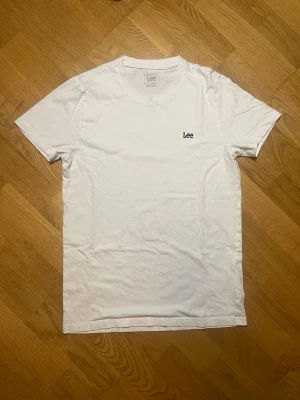 Vit t-shirt från Lee - Säljer denna klassiska vita t-shirt från Lee i storlek S (men kan även passa M) med liten svart logga broderad på bröstet. T-shirten har rund halsringning och korta ärmar. Tillverkad i mjuk bomull för en skön och enkel stil. Vid frågor, extra bilder eller liknande är det bara att kontakta mig!