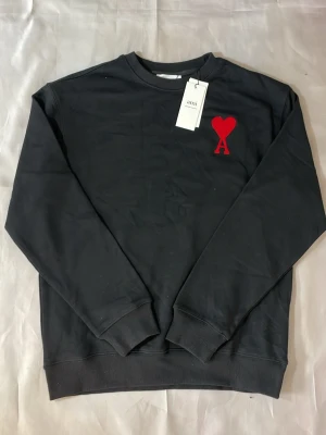 Svart sweatshirt från Ami Paris - Stilren svart sweatshirt från Ami Paris med det ikoniska röda hjärtat och bokstaven A broderat på bröstet. Tröjan har rund halsringning, ribbade muddar och är tillverkad i mjuk bomull. Perfekt för en clean och trendig look.