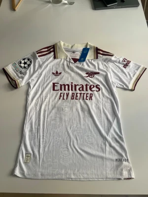 Helt ny! Arsenal 25/26 tredje Gyökeres - Snygg vit Arsenal matchtröja från Adidas med vinröda detaljer och klubbmärke på bröstet. Tröjan har trycket 'Emirates Fly Better' fram och 'GYÖKERES 14' bak. Champions League-märke på ärmen och HEAT.RDY-material för extra komfort.