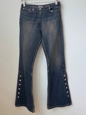Blå utsvängda jeans med knappar - Säljer ett par blå utsvängda jeans från Bisco med coola knappar längs bensluten. Jeansen har klassisk femficksmodell och en snygg tvättad look. Perfekta för dig som gillar retrostil och vill sticka ut lite extra.