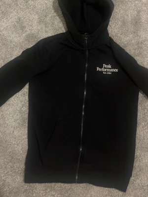 Svart hoodie Peak Performance - Snygg svart hoodie från Peak Performance med dragkedja framtill och huva. Logga i vitt på bröstet och praktisk ficka på magen. Tillverkad i mjukt material som känns skönt mot huden. Perfekt för en chill och sportig look.