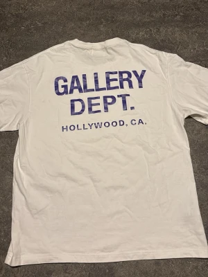 Vit Gallery Dept. t-shirt - Vit t-shirt från Gallery Dept. med tryck i blått både fram och bak. Klassisk passform med rund hals och korta ärmar. Stort tryck på ryggen med texten 'GALLERY DEPT. HOLLYWOOD, CA.' och mindre logga framtill. Perfekt för en clean och avslappnad stil.