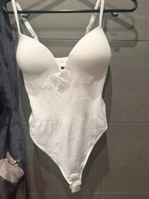 Vit vadderad body - Säljer en vit body med vadderade kupor och smala justerbara axelband. Bodyn har en djup urringning och är tillverkad i ett stretchigt material som sitter tight mot kroppen. Perfekt för att skapa en snygg siluett under kläder.