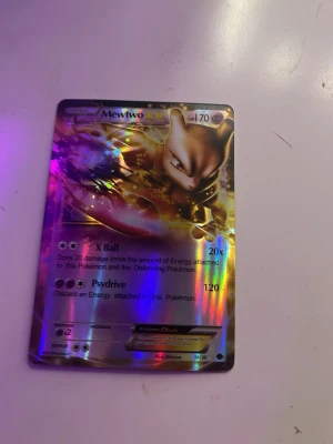 Mewtwo EX Pokémonkort holografiskt - Säljer ett holografiskt Mewtwo EX Pokémonkort med nivå 70. Kortet har en glansig yta med färger i guld, lila och blått. Motivet visar Mewtwo i en kraftfull pose och kortet har attacker som X Ball och Psydrive. Perfekt för samlare eller Pokémonfans.