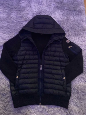 Moncler cardigan  - Snygg svart hybridjacka med quiltad pufferfront och huva. Ärmar och rygg är i mjukt stickat material, medan framsidan är vadderad för extra värme. Jackan har dragkedja framtill, två fickor med tryckknappar och en liten ficka med dragkedja på ärmen.