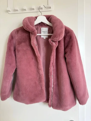 Rosa fuskpälsjacka från Name It - Säljer en fluffig rosa fuskpälsjacka från Name It med hög krage och mjukt innerfoder. Jackan har dragkedja framtill och är perfekt för kalla dagar. Trendig och mysig modell som håller dig varm hela vintern.