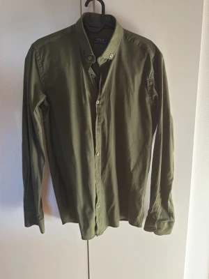 Olivgrön skjorta från Polo Ralph Lauren - Stilren olivgrön skjorta från Polo Ralph Lauren i bomull. Klassisk button-down krage och diskret logga broderad på bröstet. Långärmad modell med knappar framtill och vid manschetterna. Perfekt för en avslappnad men snygg look.