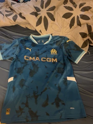 Blå Marseille matchtröja  - Snygg blå matchtröja från Olympique de Marseille med guldfärgade detaljer och klubbmärke på bröstet. Tröjan har korta ärmar, rund hals och är tillverkad i ett lätt funktionsmaterial. Perfekt för fotbollsträning eller att bära till vardags.