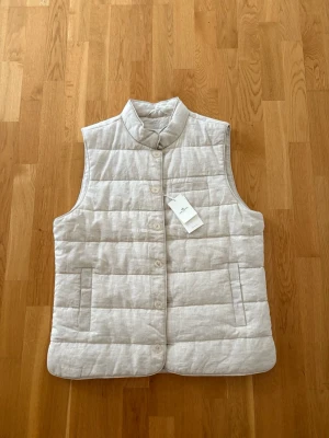 Ljusbeige quiltad dunväs - Stilren ljusbeige dunväst med ståkrage och knäppning framtill. Västen har två stora fickor på framsidan och ett quiltat mönster som ger en modern look. Perfekt att slänga över en hoodie eller tröja för extra värme. Bara testad, lapp kvar.