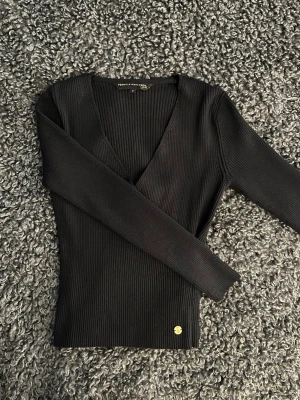 Svart ribbad v-ringad topp - Snygg svart ribbad topp med v-ringning och långa ärmar. Tillverkad i stretchigt material som sitter tight och markerar figuren. Perfekt att styla med jeans eller kjol för en clean look.