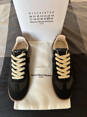 Maison Margiela svarta sneakers - Svarta sneakers från Maison Martin Margiela med beige snörning och gummisula. Skorna har en klassisk siluett med detaljer i skinn och mocka, samt diskret branding på plösen. Perfekta för dig som gillar stilrena och trendiga sneakers.