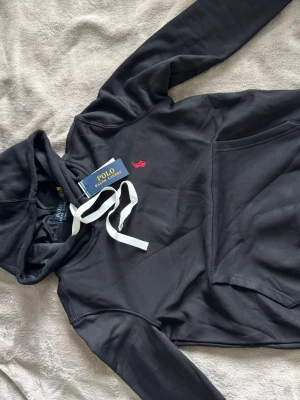 Svart hoodie från Polo Ralph Lauren - Snygg svart hoodie från Polo Ralph Lauren med klassisk röd logga broderad på bröstet. Hoodien har vit dragsko i huvan och en stor magficka framtill. Tillverkad i mjukt material som känns skönt mot huden. Perfekt för en avslappnad och trendig look. Storlek S