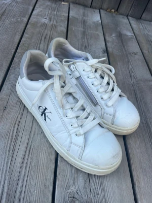 Vita Calvin Klein sneakers med dragkedja - Jättefina vita skor från Calvin Klein jeans med en fin silvrig detalj där bak! Lite slitna längst fram, på snörena och på sulan men annars bra skick🩵säljer då de tyvärr inte kommer till användning längre. Skriv om ni har frågor eller vill ha fler bilder på skorna så fixar jag det så fort jag kan! Pris kan diskuteras.