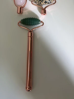 Ansiktsroller med utbytbara huvuden - Snygg ansiktsroller i roséguld med tre utbytbara huvuden: ett i grön sten med räfflad yta, ett i rosa sten och ett i vit sten med svarta prickar. Perfekt för ansiktsmassage och hudvård. Handtaget är i metall och ger ett lyxigt intryck.