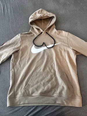 Beige Nike hoodie med logga - Snygg beige hoodie från Nike med stor vit logga på bröstet och svarta snören i huvan. Tröjan har en klassisk känguruficka framtill och svarta samt röda detaljer längs ärmarna. Perfekt för en avslappnad streetwear-look.