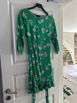 Grön blommig klänning med volang - Grön klänning med blommigt mönster och volangkant nertill. Klänningen har trekvartsärm och knytband i midjan. Perfekt för dig som gillar färg och mönster. Materialet känns mjukt och lätt, troligen polyester.