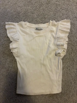 Vit ribbad topp med volangärmar Zara - Supersöt vit ribbad topp från Zara med volangdetaljer på ärmarna. Toppen har en rund halsringning och är tillverkad i ett mjukt, stretchigt material som sitter snyggt på kroppen. Perfekt att styla med jeans eller kjol.