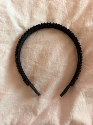 Svart diadem med glittrande stenar - Snyggt svart diadem dekorerat med glittrande svarta stenar som ger en lyxig känsla. Diademet har en klassisk form och är täckt av små, facetterade pärlor som reflekterar ljuset. Perfekt accessoar för att lyfta din look och ge extra edge till frisyren.