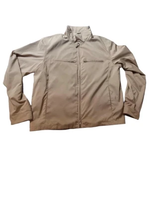 Beige vindjacka från Loro Piana - Stilren beige vindjacka från Loro Piana med hög krage och två bröstfickor med dragkedja. Jackan har hel dragkedja framtill och är tillverkad i ett lätt, vindtätt material. Perfekt för dig som vill ha en clean och klassisk look.
