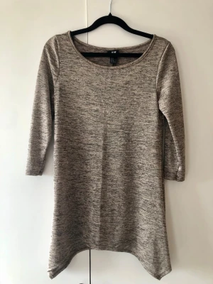 H&M Glittrig Tröja / Topp Melerad m. Flikar – Stl XS - Fin och stretchig tröja från H&M i glittrigt melerat tyg. Modell med trekvartsärm, rund hals och snyggt asymmetrisk fåll som hänger ned i flikar i sidorna. Perfekt till både vardag och fest. Storlek: XS Mått: Axelbredd ca 35 cm, längd ca 70 cm Material: 95% polyester, 5% elastan Skick: Mycket gott skick