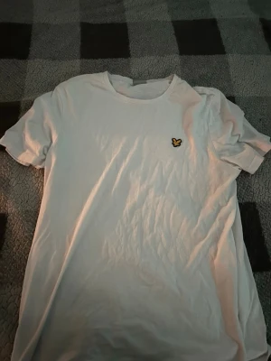 Vit t-shirt med gul logga - Säljer en enkel vit t-shirt med rund hals och en liten gul broderad logga på bröstet. T-shirten har korta ärmar och är gjord i mjuk bomull, perfekt för en clean och stilren look.