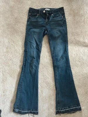 Blå bootcut jeans med rå kant - Säljer ett par blå bootcut jeans med rå, fransad kant nedtill. Jeansen har klassisk femficksmodell, knappgylf och lätt slitna detaljer framtill. Materialet är denim i bomull med lite stretch för skön passform.