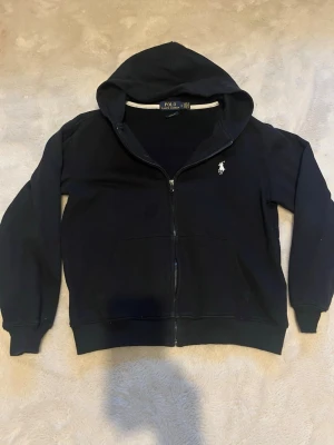 Svart hoodie från Polo Ralph Lauren - Svart hoodie med dragkedja från Polo Ralph Lauren. Klassisk design med huva, fickor framtill och vit broderad logga på bröstet. Tillverkad i mjukt bomullsmaterial som är skönt att bära. Perfekt för en avslappnad och stilren look. Liten i storlek. 