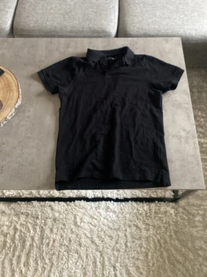 Svart kortärmad polotopp - En stilren svart polotopp med klassisk krage och korta ärmar. Perfekt till jeans eller kjol för en enkel och clean look. Materialet känns mjukt och bekvämt mot huden.