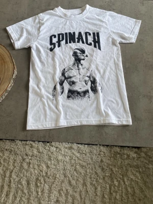 Vit t-shirt med Popeye-tryck - Säljer en vit t-shirt med stort svart tryck av Popeye och texten 'SPINACH' på bröstet. Klassisk rund hals och korta ärmar. Perfekt för dig som gillar coola och unika motiv på dina kläder.