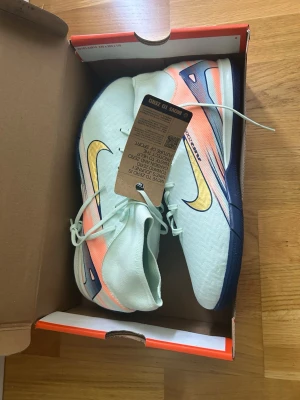 Nike fotbollsskor ljusblå/gul - Snygga fotbollsskor från Nike i ljusblått med gula och orange detaljer samt mörkblå sula. Skorna har hög krage för extra stöd runt ankeln och klassisk snörning. Tillverkade i syntetmaterial för bra grepp och känsla på planen.