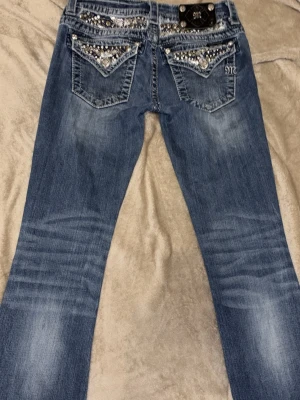Miss Me Jeans - Defekt på femte bilden!! Innerben: 72cm Midjemått: 36-37cm. Säljer dyrt eftersom jag tkr verkligen om dem. Snygga blå bootcut jeans med slitningar och dekorativa detaljer på fickorna. Jeansen har låg midja, klassisk femficksmodell och markerade sömmar. Perfekta för dig som gillar en avslappnad men trendig stil.