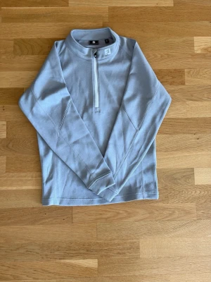 Grå halvzip tröja från Foot joy - Snygg ljusblå långärmad tröja från Foot joy med halv dragkedja framtill och hög krage. Perfekt för lager-på-lager och har en sportig look. Tillverkad i mjukt material som känns skönt mot huden. Stilrena sömmar och minimalistisk design. Perfekt för golf eller liknande aktiviteter