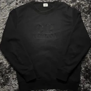 C.P. Company Tröja - Klassisk svart C.P. Company-sweatshirt med broderad logga framtill. Storlek L, men passar även M beroende på önskad passform. Stilren, lättmatchad och perfekt till vardags/streetwear. QR-kod finns som äkthetsbevis. Skick enligt bilder.