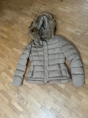 Beige pufferjacka med fuskpäls - Snygg beige pufferjacka från So Sweet med stor huva och fuskpälsdetalj. Jackan har dragkedja och knappar framtill samt två fickor med dragkedja. Perfekt för kalla vinterdagar och ger en trendig look. Priset kan diskuteras 