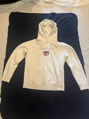 Vit hoodie från GANT med logga - Snygg vit hoodie från GANT med stor broderad logga och sköld på bröstet. Klassisk känguruficka framtill och huva med dragsko. Tillverkad i mjukt material som känns skönt mot huden. Perfekt för en avslappnad och stilren look.