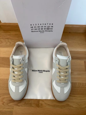Maison Margiela sneakers vita/grå - Maison Margiela sneakers i vitt och ljusgrått läder och mocka, med beige snörning och klassisk gummisula. Snygg retrostil med låg profil och diskreta detaljer. Perfekta för dig som gillar minimalistisk och stilren design.