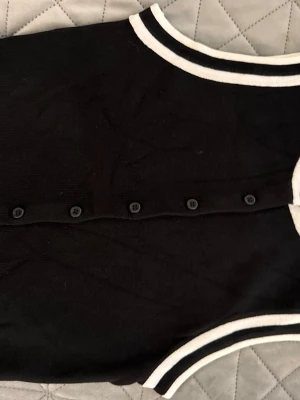 Svart kofta med vita ränder - Snygg svart kofta med vita ränder vid krage och ärmslut. Knäppning framtill med svarta knappar. Klassisk och stilren design som passar till mycket. Mjuk och skön kvalitet.