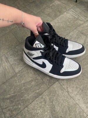 Svarta och vita Nike Air Jordan 1 Mid - Säljer ett par Nike Air Jordan 1 Mid sneakers i svart och vitt. Klassisk design med svart swoosh, perforerad tåbox och högt skaft. Skorna har snörning och är tillverkade i en kombination av läder och textil. Perfekta för dig som gillar streetwear och ikoniska sneakers.