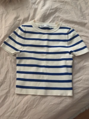 Randig topp från Zara i vitt och blått - Kortärmad topp från Zara i vitt med blå horisontella ränder. Klassisk rund halsringning och rak passform. Perfekt för en avslappnad och stilren look. Materialet känns mjukt och bekvämt mot huden.