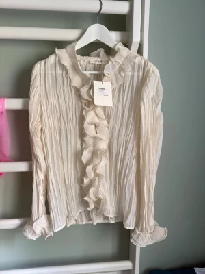 Vit/beige plisserad blus med volang - Supersöt blus i plisserat tyg med långa ärmar och volangdetaljer längs krage och knappslå. Blusen har en romantisk känsla med ryschade muddar och lätt transparent material. Perfekt att styla med jeans eller kjol för en trendig look.