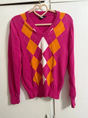 Rosa stickad tröja med argylemönster - Snygg rosa stickad tröja med V-ringning och färgglatt argylemönster i orange, vitt och rosa på framsidan. Tröjan har långa ärmar, ribbade muddar och är gjord i ett mjukt, tunt material. Perfekt för dig som gillar färg och mönster!