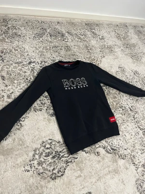 Svart sweatshirt från Hugo Boss - Snygg svart sweatshirt från Hugo Boss med vit logga på bröstet och en röd detalj vid nederkanten. Tröjan har rund halsringning och långa ärmar. Perfekt för en stilren och avslappnad look.