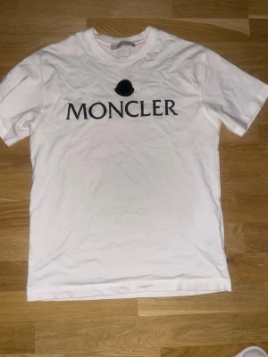 Vit Moncler t-shirt med logga - Säljer en stilren vit t-shirt från Moncler med stor svart logga och text på bröstet. Klassisk rund halsringning och korta ärmar. Perfekt till jeans eller shorts för en clean look.