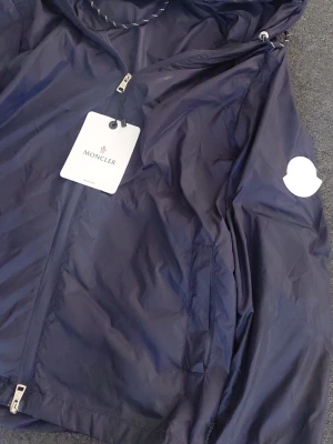 Mörkblå vindjacka från Moncler - Snygg mörkblå vindjacka från Moncler med huva och dragkedja framtill. Jackan har två sidofickor med dragkedja och en vit Moncler-logga på ärmen. Tillverkad i lätt, glansigt material som står emot vind. Perfekt för dig som vill ha en stilren och sportig look.