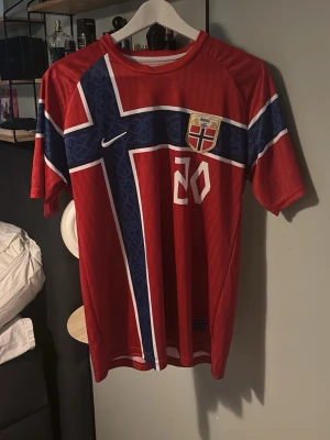 Norge fotbollströja Nusa 20 - Röd Norge landslagströja med blått och vitt kors, nummer 20 och namnet Nusa på ryggen. Tröjan har korta ärmar och är tillverkad i ett lätt och ventilerande material, perfekt för fotbollsträning eller match.