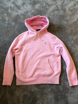 Rosa hoodie från Polo Ralph Lauren -  Köpt på Johnells för 3 veckor sedan. Säljer en ljusrosa hoodie från Polo Ralph Lauren med klassisk huva och dragsnören.  Tröjan har en känguruficka framtill och den ikoniska blå loggan broderad på bröstet. Tillverkad i mjukt bomullsmaterial som är riktigt skönt att ha på sig.