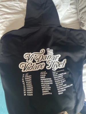 Hov1 hoodie - Säljer en svart hoodie med stor vit text på ryggen 'Vi Danar Vidare Livet' och turnélista. På bröstet finns en liten broderad krona. Hoodien har huva och magficka. Perfekt för dig som gillar unik streetwear med festivalvibbar.