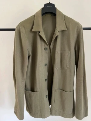 Olivgrön chorejacket - Stilren olivgrön overshirt med tre framfickor och knäppning framtill. Skjortkrage och raka ärmar ger en avslappnad look. Tillverkad i ett mjukt, vävt material som passar perfekt för lager på lager. Enkel och clean design utan synliga loggor.